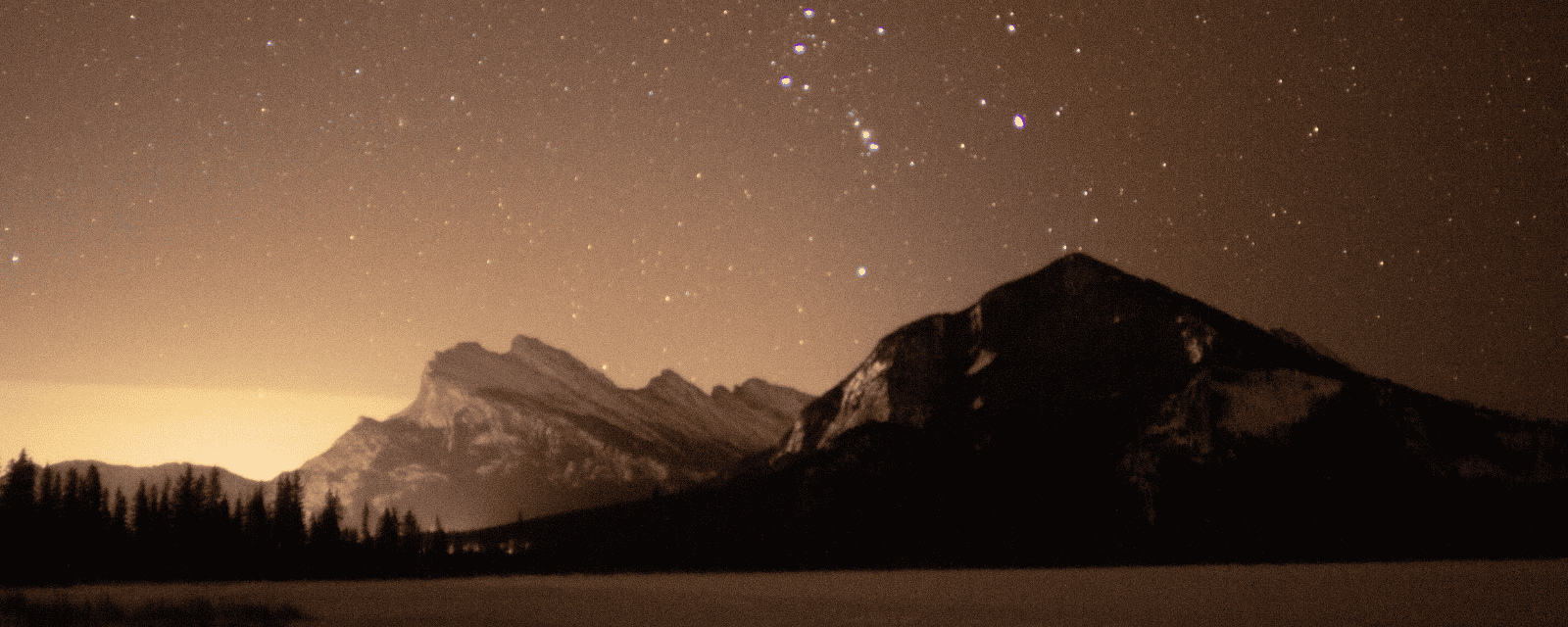 A starry night sky over dark mountain peaks, Orion rising above the ridgeline