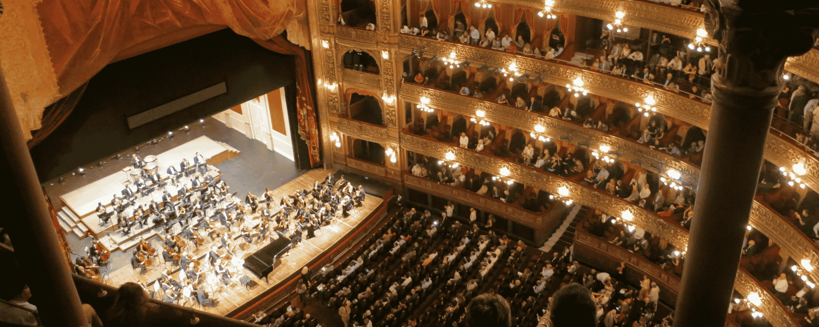 A grand gilded opera house seen from above, an orchestra on stage and the audience filling every balcony